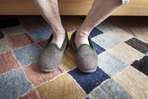 Pflege zu Hause Ein alter Mann sitzt auf der Kante seines Bettes, man sieht nur seine nackten Unterschenkel, die Füße stecken in beigen Filzpantoffeln.