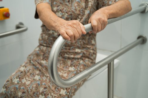 Barrierefreies Badezimmer Eine ältere Frau stzt auf einem geschlossenen Toilettendeckel, sie hält sich mit beiden Händen an einem Haltegriff fest, der neben der Toilette angebracht ist.