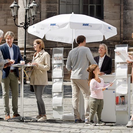 Ein VdK-Infostand in einer Fußgängerzone, eine Frau spricht mit einem Passanten, am Stehtisch daneben unterhalten sich Menschen. 