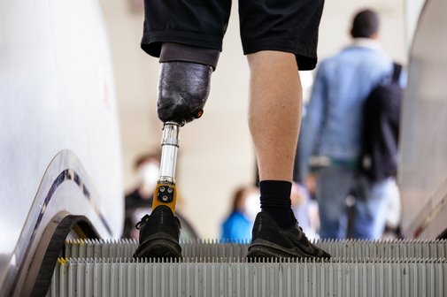 Ein Mann mit einer Beinprothese f&auml;hrt Rolltreppe. Man sieht nur seine F&uuml;&szlig;e und Waden, an einem Bein hat er eine Unterschenkelprothese.