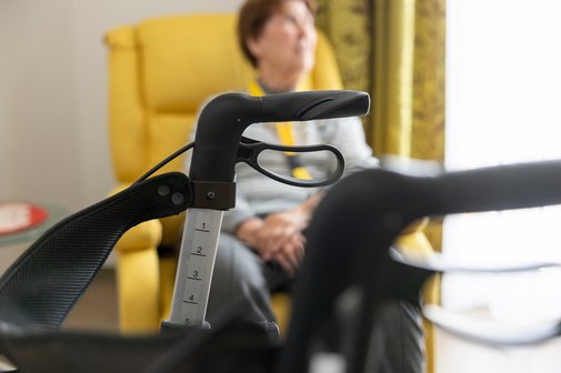 Eine &auml;ltere Frau sitzt in einem gelbem Sessel. Im Vordergrund steht ein Rollator.