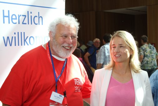 Verena Bentele mit weißem Sakko und rosa Shirt neben einem großen, kräftigen Mann mit weißem Haar und Bart in knallrotem Shirt, der vor Freude strahlt. 