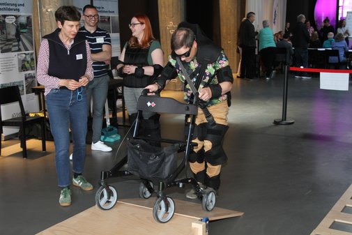Mehrere Menschen bei Gehversuchen mit Rollator und Alterssimulationsanzug