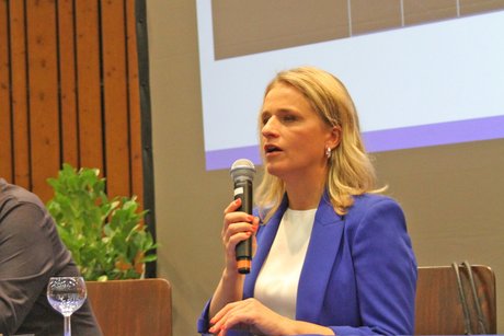 Frau mit blondem Haar und blauem Sakko steht vor dem Saal und spricht in ein Mikro. 