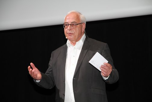 Mann im weißen Hemd und dunklem Sakko und Zettel mit Stichpunkten für seine Rede in der linken Hand. 