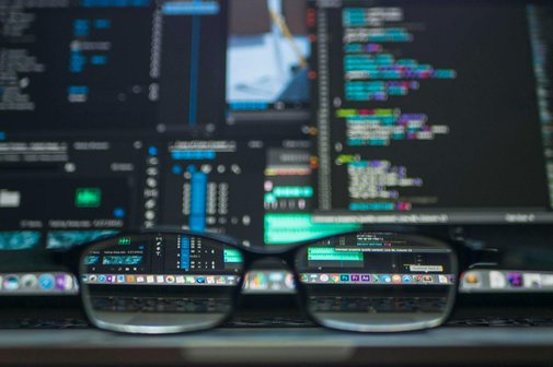 Im Hintergrund mehrere Bildschirme, über die Datenkolonnen laufen. Vorne eine Brille, durch deren Gläser die verschwommenen Daten scharf und lesbar werden. 