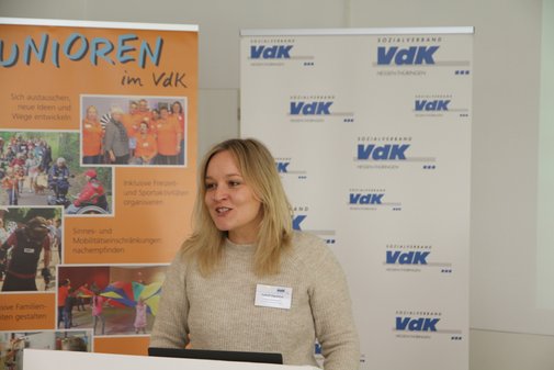 Junge Frau mit blondem mittellangen Haar in einem beigen Pullover am Rednerpult