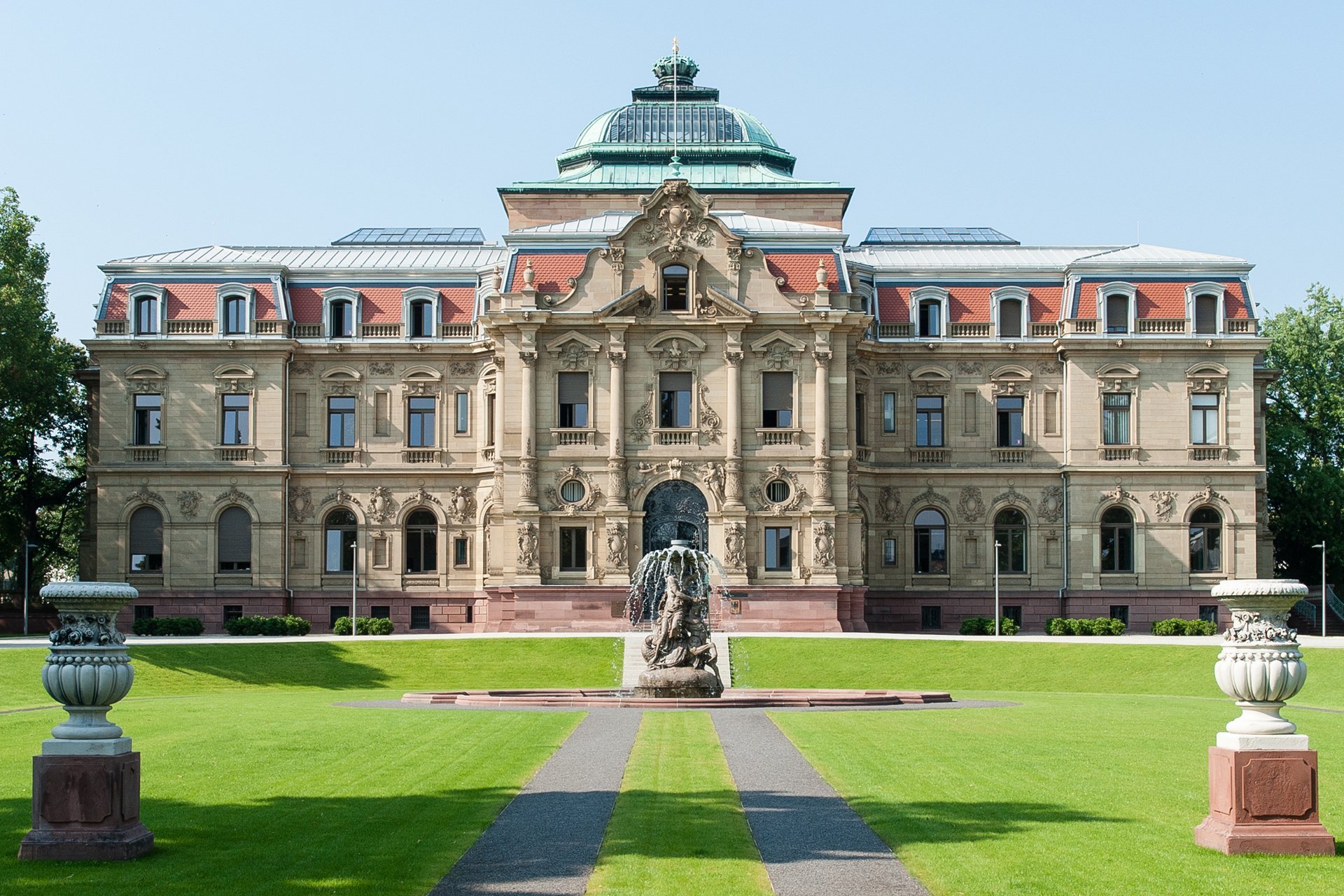 Blick auf einen im Stil des Neobarocks gestaltetes Palastgeb&auml;ude im Sonnenschein mit gro&szlig;en Rasenfl&auml;chen und einem Brunnen davor.