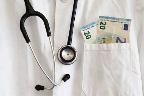 Nahaufnahme eines Arztkittels. Man sieht nur den Brustbereich und ein um den Hals gehängtes Stethoskop. Aus der Brusttasche des Kittels schauen zwei 20-Euro-Scheine heraus.