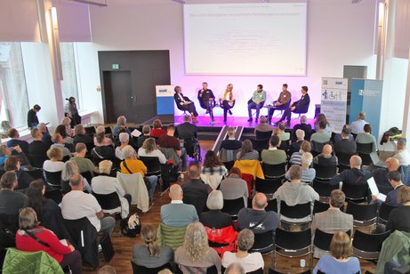 Ein Blick von oben in einen Vortragsraum. Der Raum ist gut gef&uuml;llt mit etwa 50 G&auml;sten. Vorne ist eine kleine B&uuml;hne, auf der 6 Menschen sitzen, die an einer Podiumsdiskussion teilnehmen.