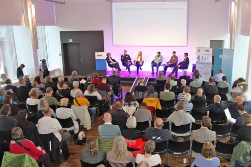 Ein Blick von oben in einen Vortragsraum. Der Raum ist gut gefüllt mit etwa 50 Gästen. Vorne ist eine kleine Bühne, auf der 6 Menschen sitzen, die an einer Podiumsdiskussion teilnehmen.