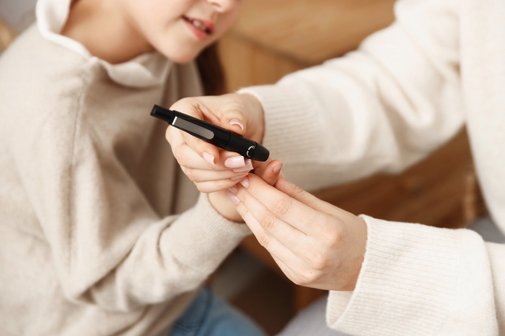 Eine Frau in hellem Pullover misse den Blutzucker eines Kindes in einem beigefarbenen Pullover