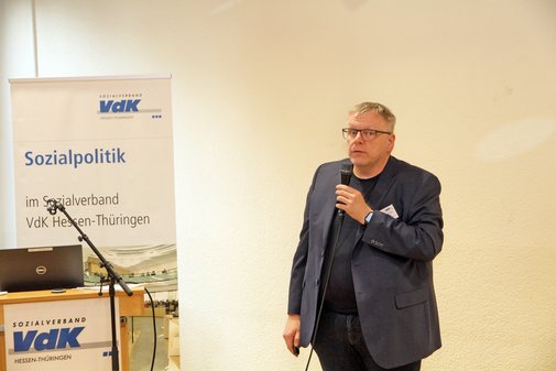 Ein Mann steht vor einer Wand und spricht in ein Mikrofon, neben ihm en Rednerpult und ein Roll-up des Sozialverbands VdK Hessen-Th&uuml;ringen
