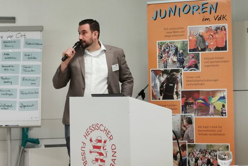 Mann mit dunklem Haar und Bart in braunem Jacket mit wei&szlig;em Hemd spricht in Mikrofon