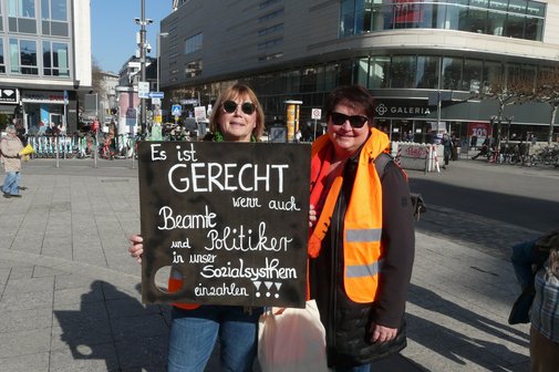 An der Hauptwache in Frankfurt halten bei strahlendem Sonnenschein zwei Frauen mit Sonnenbrillen ein Schild mit der Beschriftung "Es ist gerecht, wenn auch Beamte und Politiker in unser Sozialsystem einzahlen!" hoch.