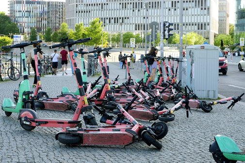 Mehrere E-Scooter, größtenteils umgekippt, liegen und stehen chaotisch auf einem gepflasterten Gehweg in einer Stadt. Im Hintergrund sind moderne Bürogebäude, Fußgänger sowie parkende Fahrräder zu sehen. Die E-Scooter gehören verschiedenen Anbietern und blockieren teilweise den Weg