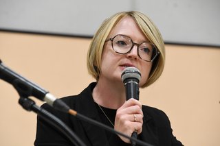 Frau mit blondem Pagenkopf und Brille hält sich das Mikro sehr nahe an den Mund, sagt etwas und wirkt dabei sehr ernst und besorgt. 