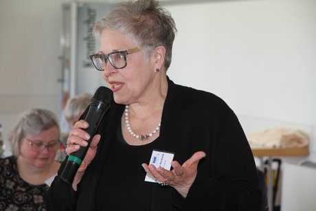 Frau in Schwarz blickt nach links aus dem Bild heraus und hat die freie Hand, die nicht das Mikro h&auml;lt, zur gestischen Bekr&auml;ftigung ihrer Argumentation in Brusth&ouml;he nach oben ge&ouml;ffnet und die Finger gespreizt. 