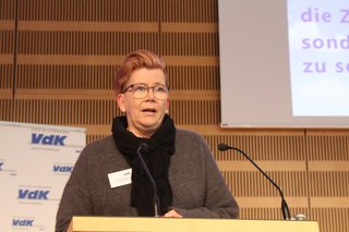 Frau mit schwarzem Schal spricht zu Publikum 