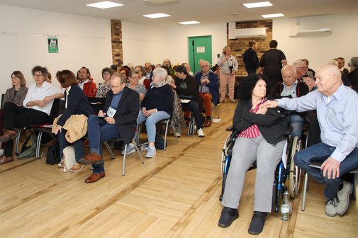 Blick ins Publikum bei einer Vortragsveranstaltung. Der Raum ist vollbesetzt. Im Vordergrund hält ein Mann die Hand seiner Frau, die im Rollstuhl sitzt.