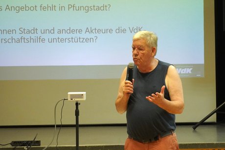 &Auml;lterer Mann in Tanktop und Shorts steht mit dem Mikro in der Hand am Kopf des Saals und spricht engagiert zu den Zuh&ouml;rern. 