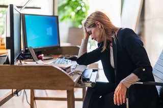 Eine erschöpfte Frau sitzt am Schreibtisch vor ihrem Computer und hat den Kopf aufgestützt.