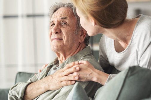 Vater und Tochter Älterer Mann sitzt auf der Couch, eine jüngere Frau umarmt ihn von hinten.