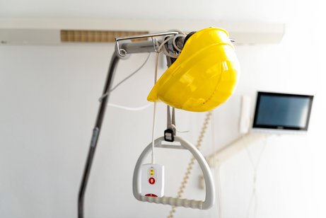 Ein gelber Bauhelm h&auml;ngt an einem Haltegriff &uuml;ber einem Krankenhausbett; im Hintergrund sind eine Klinikwand und ein Bildschirm zu sehen.