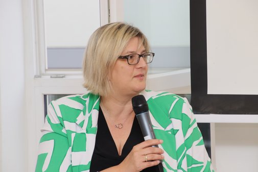 Frau mit blondem Haar und Brille und einem Blazer mit auffälligem grün-weißen Blumendekor spricht in ein Mikro. 