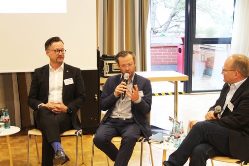 Drei Personen sitzen auf St&uuml;hlen. Einer h&auml;lt ein Mikrophon in der Hand und gestikuliert mit der anderen Hand.