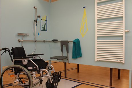 Bad in der Ausstellung Hallo Freiheit! Barrierefreies Badezimmer mit Duschbereich, Haltegriffen und Duschsitz; daneben steht ein Rollstuhl, an der Wand hängt ein Handtuch und ein Heizkörper ist zu sehen