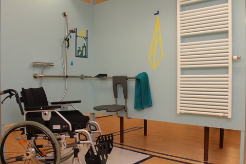 Barrierefreies Badezimmer mit Duschbereich, Haltegriffen und Duschsitz; daneben steht ein Rollstuhl, an der Wand h&auml;ngt ein Handtuch und ein Heizk&ouml;rper ist zu sehen