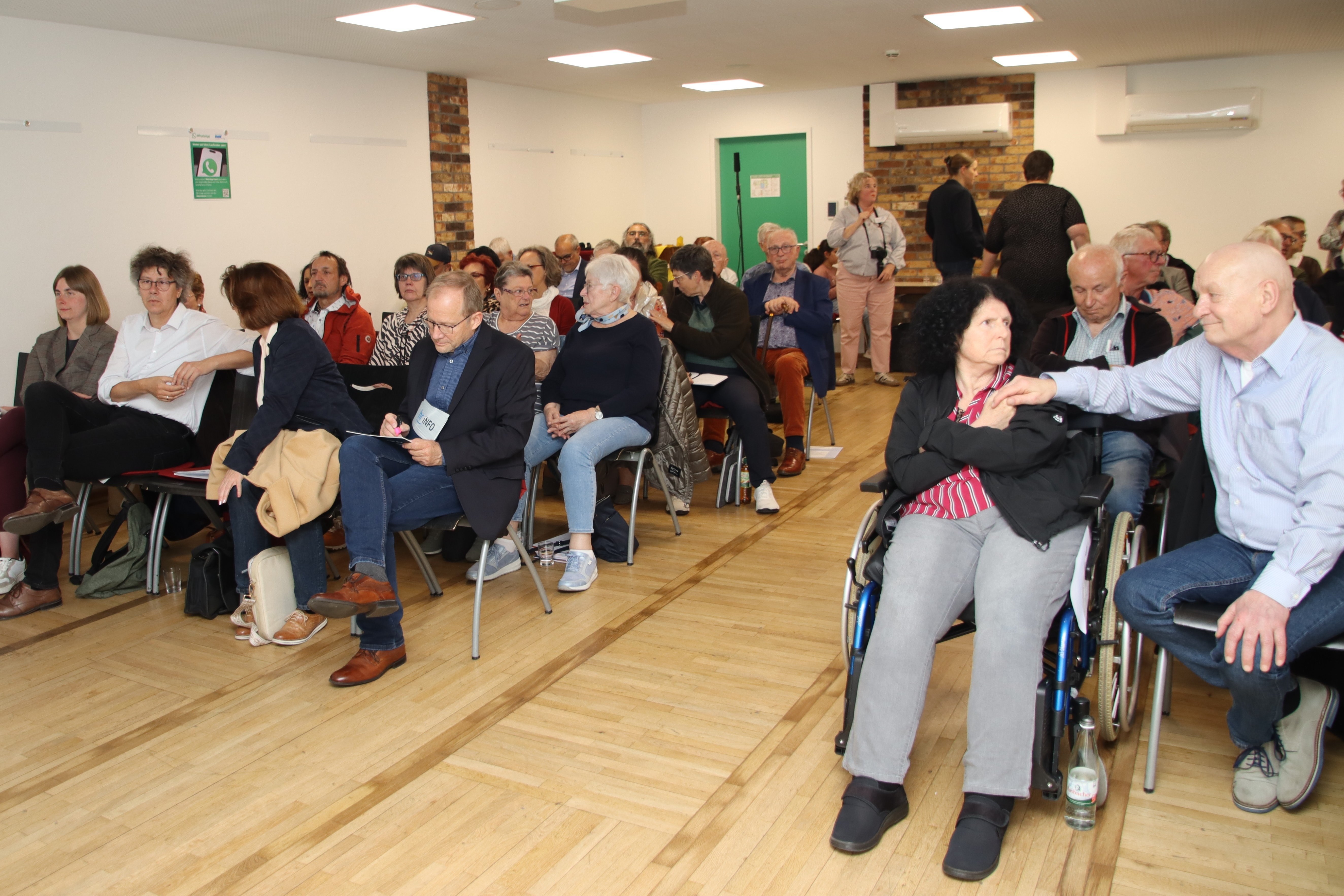 Blick ins Publikum bei einer Vortragsveranstaltung. Der Raum ist vollbesetzt. Im Vordergrund hält ein Mann die Hand seiner Frau, die im Rollstuhl sitzt.