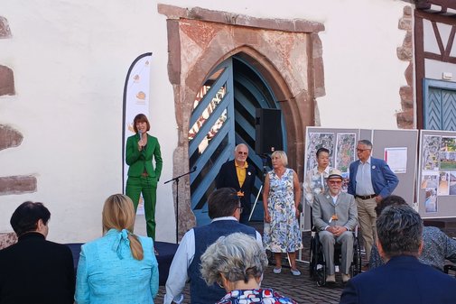 Vor einem Haus spricht eine Frau in g&uuml;nem Hosenanzug in ein Mikro
