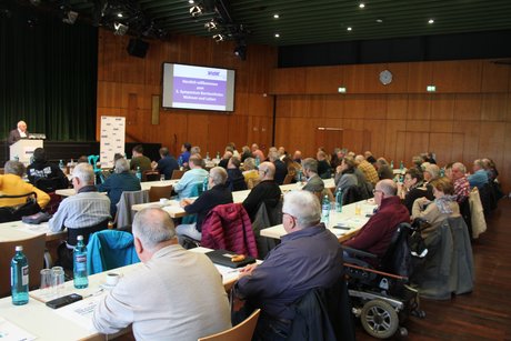 Blick auf das Publikum Blick in einen großen holzgetäfelten Saal, an dem an langen Tischen viele Menschen sitzen und einem Vortragsredner am Pult zuhören.