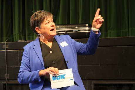 Katja Lüke, Moderatorin des Symposiums Frau im Rollstuhl erklärt etwas und zeigt mit erhobenem Zeigefinger auf eine Leinwand, die allerdings nicht im Bild zu sehen ist.