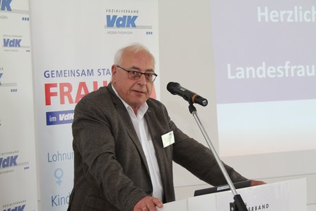 Mann im dunklen Sakko und wei&szlig;em Hemd st&uuml;tzt sich beim Reden auf das Stehpult auf. 