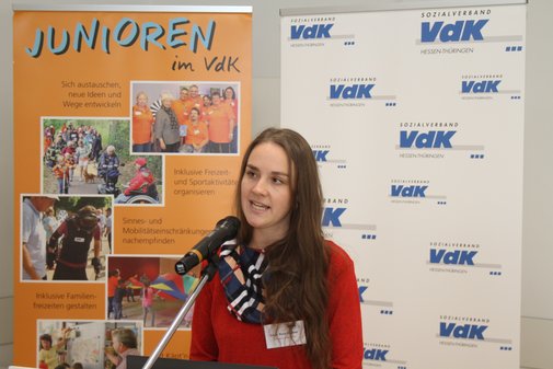 Eine Frau mit langem braunem Haar und rotem Pullover an einem Rednerpult