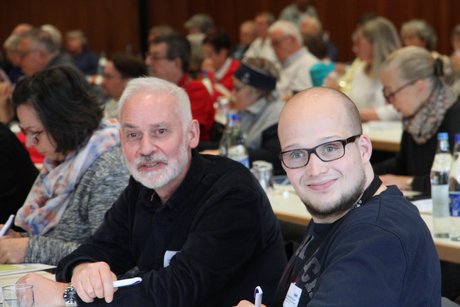 Ein älterer weißhaariger Mann und ein junger Mann mit Brille und Glatze lächeln in die Kamera.