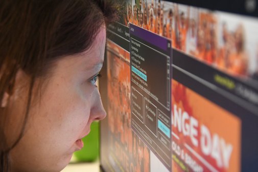 Eine Frau blickt aus nächster Nähe auf einen Computerbildschirm. Ihr Gesicht ist sehr nah an der Bildschirmoberfläche, auf der mehrere Fenster mit Text und bunten Bildern zu sehen sind. Im Hintergrund ist ein verschwommenes Gruppenfoto von Menschen in oranger Kleidung sowie der Schriftzug „ORANGE DAY“ erkennbar. Die Szene deutet auf eine eingeschränkte Sehfähigkeit der Frau hin.