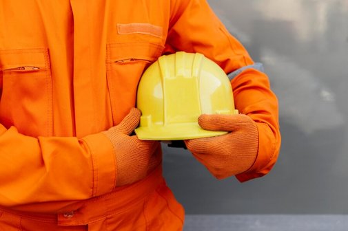 Ein Mensch in einem orangefarbenen Overall tr&auml;gt einen gelben Helm in den H&auml;nden