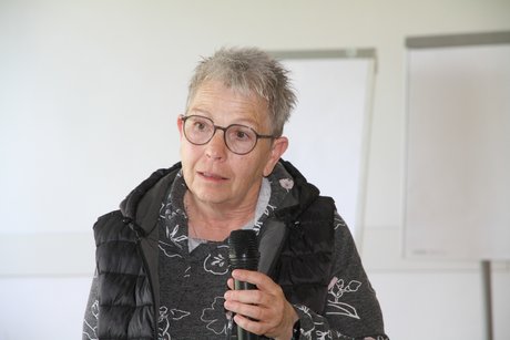 Frau mit Brille und grauem Kurzhaarschnitt blickt nachdenklich und fragend in die Kamera, w&auml;hrend sie etwas ins Mikro zu sagen scheint. 