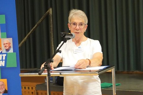 Frau mit Kurzhaarschnitt und Brille in wei&szlig;er Bluse steht an einem hohen Rednerpult und blickt direkt ins Publikum. 