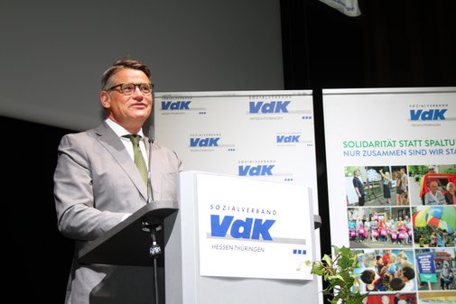 Mann mit Brille, im grauen Anzug, adressiert vom Rednerpult aus das Publikum. 