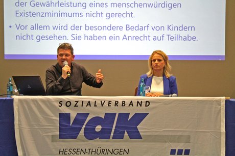 Langer Tisch mit VdK-Fahne bedeckt. Dahinter Verena Bentele, die zuhört, während Philipp Stielow etwas ins Mikro spricht.