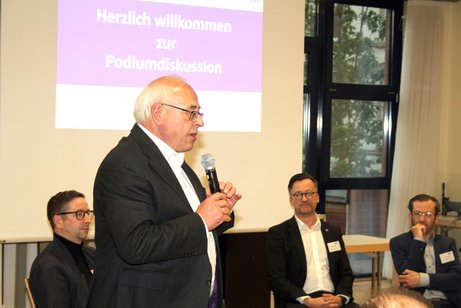 Ein Mann steht auf einer B&uuml;hne und spricht vor Publikum. Im Hintergrund sitzen die Teilnehmenden einer Podiumsdikussion.