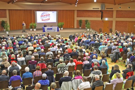 Blick über volle Stuhlreihen in einer mit Holz getäfelten großen Veranstaltungshalle. 