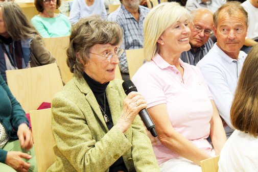 &Auml;ltere Frau sitzt im Publikum und spricht in ein Mikrofon