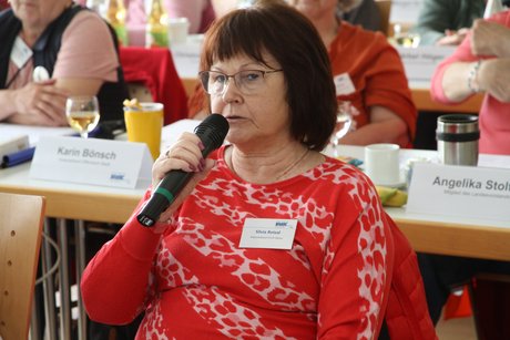 Frau mit dunklem Pagenkopf und Brille, die in einer der vorderen Tischreihen im Konferenzraum sitzt, mit einer Wortmeldung, die sie in ein Mikrofon spricht. 