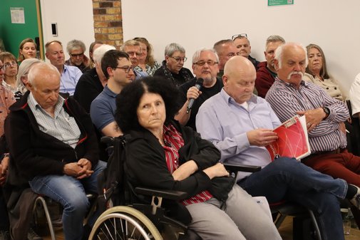 Blick über die Publikumsreihen. Im Vordergrund: Monika Schneider, im Rollstuhl. Neben ihr blättert ihr Mann Jürgen gerade in einer Broschüre. 